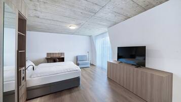 Une chambre d’hôtel moderne avec un plafond en bois, un lit, un téléviseur posé sur un meuble en bois, un bureau avec une chaise et une grande fenêtre ornée de rideaux légers.