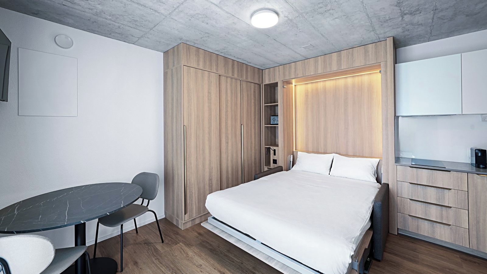 Ein modernes Hotelzimmer mit Bett, Schreibtisch und Stuhl. Ein Kleiderschrank mit Spiegel und Regalen vorhanden.