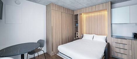 Una habitación de hotel moderna con cama, escritorio y silla. Se observa un armario con espejo y una estantería.