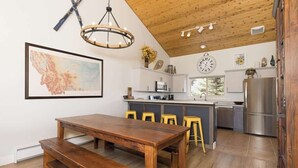Une cuisine avec un plancher en bois, une table à manger en bois avec des bancs, un lustre et une horloge au mur.