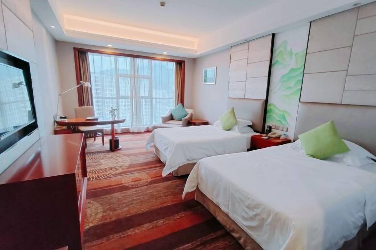 Habitación de hotel con dos camas, un escritorio, un televisor y un ventanal grande.