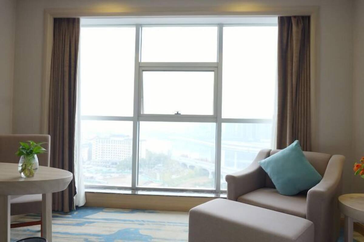 Habitación de hotel con un ventanal grande, una mesa redonda, un sofá y una mesita con una planta.
