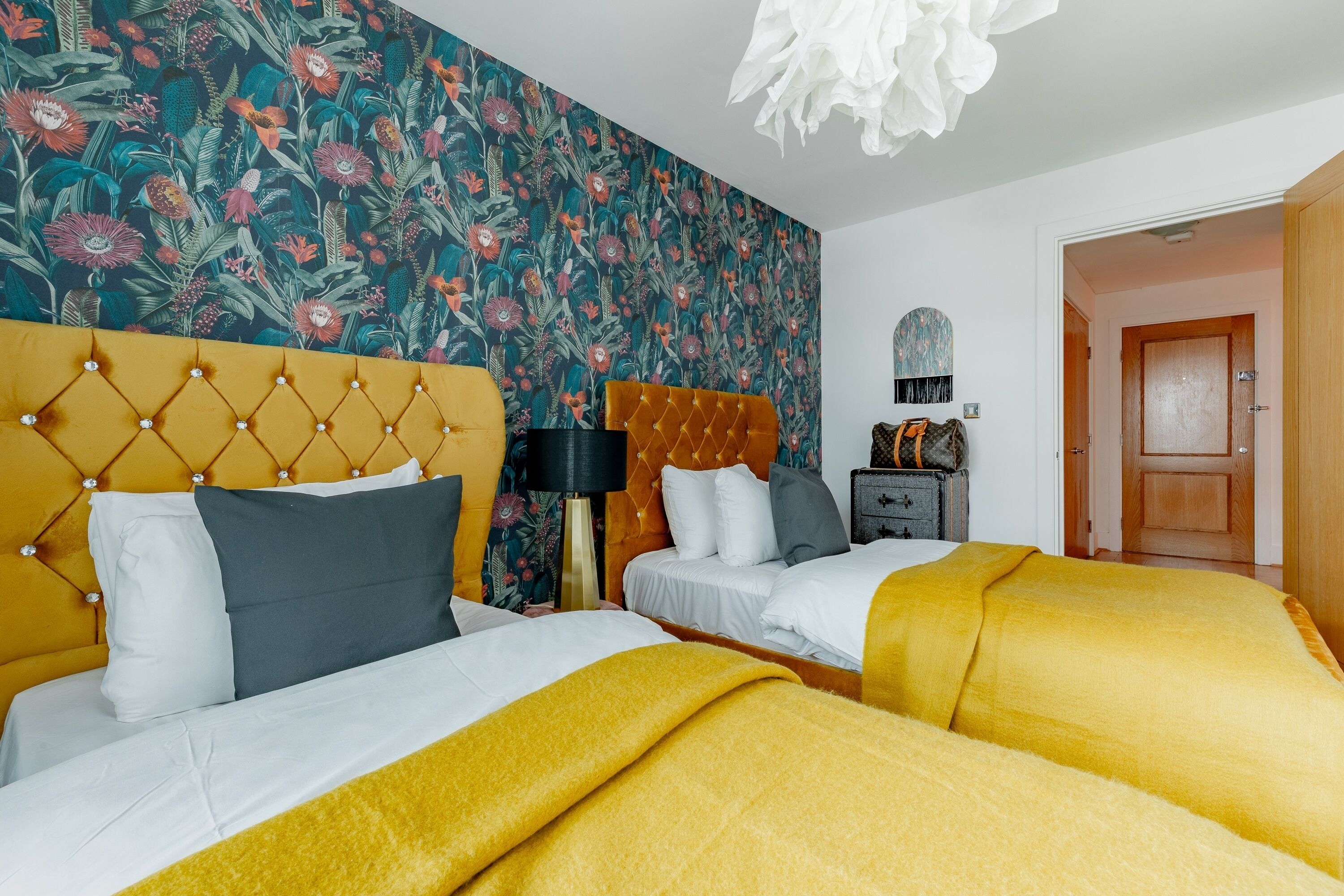 Ein Hotelzimmer mit zwei Betten, floralem Wandpapier, einem gepolsterten Kopfteil, einer Holztür und einem Nachttisch mit Lampe.
