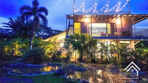 Uma villa moderna com varanda, cercada por vegetação tropical e um pequeno lago.