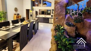 Uma sala de jantar moderna com uma mesa comprida e cadeiras, uma cozinha ao fundo e uma parede de rocha com plantas.