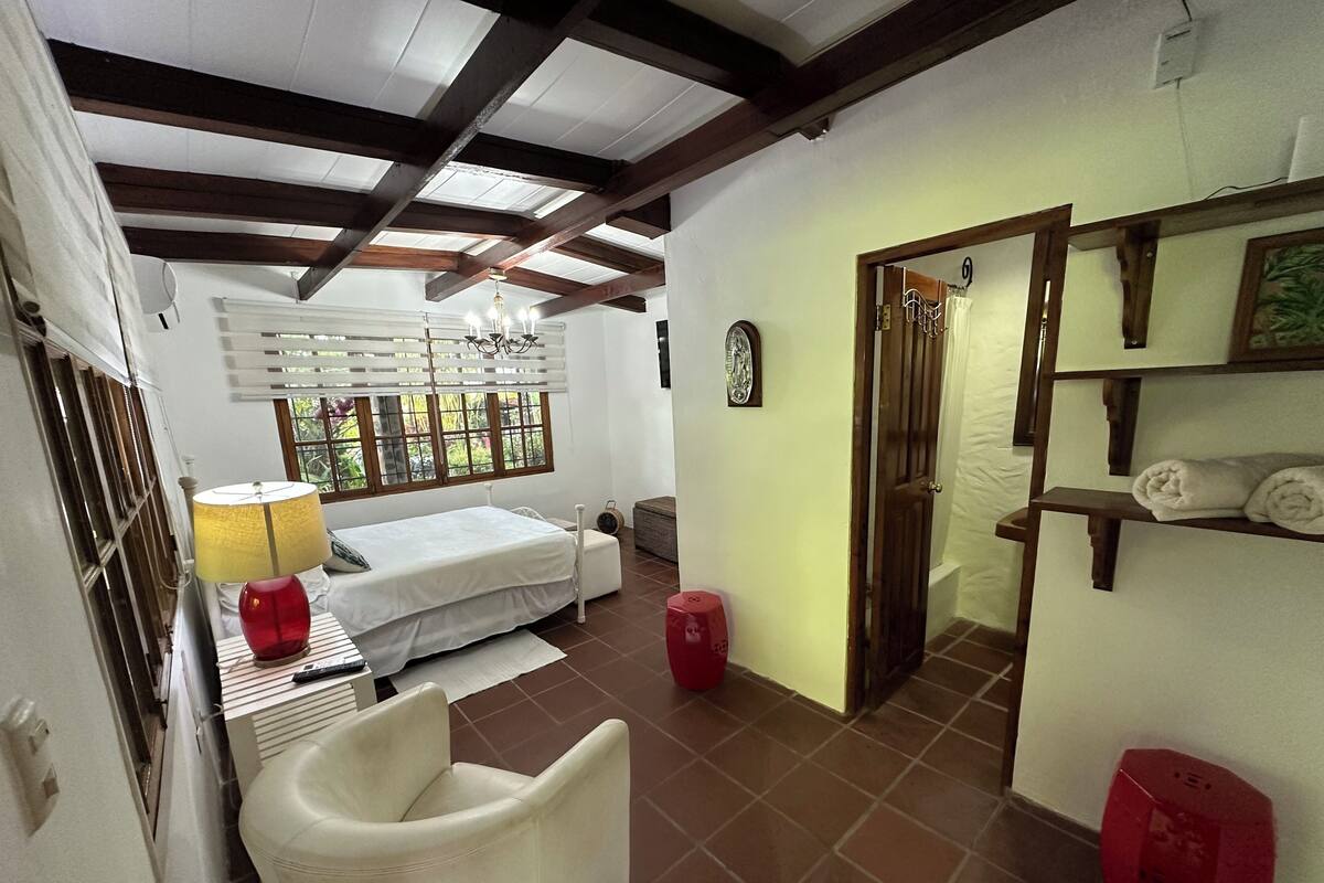 Une chambre à coucher comprenant un lit, une chaise, un tabouret rouge, une fenêtre avec des stores et une porte donnant sur une autre pièce.