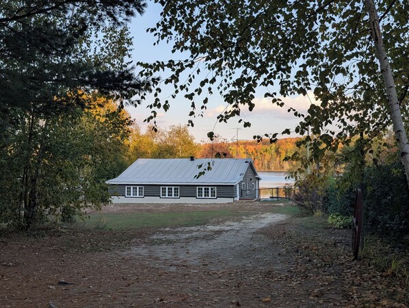A rustic cabin with a metal roof, surrounded by dense trees and a dirt path leading to it. - Charming waterfront cottage - pets welcome! (Cayamant)