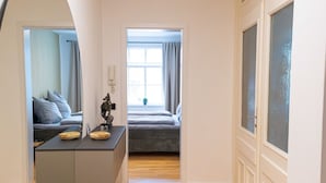 Een moderne, goed verlichte kamer met een houten vloer, een grote spiegel, een bed met een grijs hoofdeinde, een klein tafeltje met decoratieve items, en een raam met gordijnen.