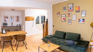 Een moderne woonkamer met een houten eettafel, een groene bank en een salontafel. Op de muur hangen ingelijste kunstwerken en er staat een houten kast met een fles wijn.