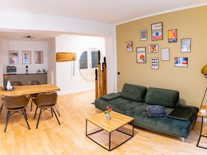 Ein modernes Wohnzimmer mit einem Holzesstisch, einem grünen Sofa und einem Couchtisch. An der Wand befinden sich gerahmte Kunstwerke und eine Holzkommode mit einer Weinflasche.