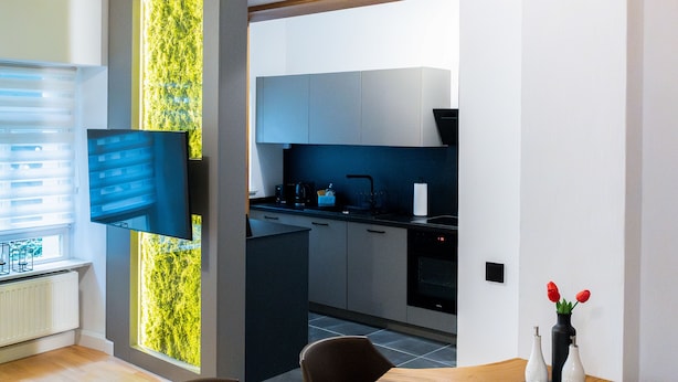 Een moderne keuken met grijze kastjes, een zwart aanrecht en een inbouwoven. Er is een eethoek met een houten tafel en stoelen, en een tv aan de muur bevestigd.