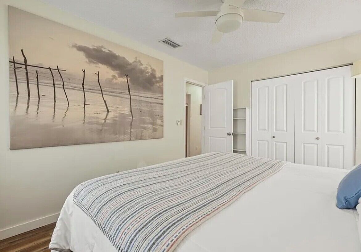 Un dormitorio con un cuadro grande de una playa, una cama con una colcha a rayas, un ventilador de techo y puertas blancas.