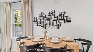 Un comedor con una mesa de madera dispuesta para cuatro, sillas negras, un jarrón con flores y una obra de arte abstracta en la pared.