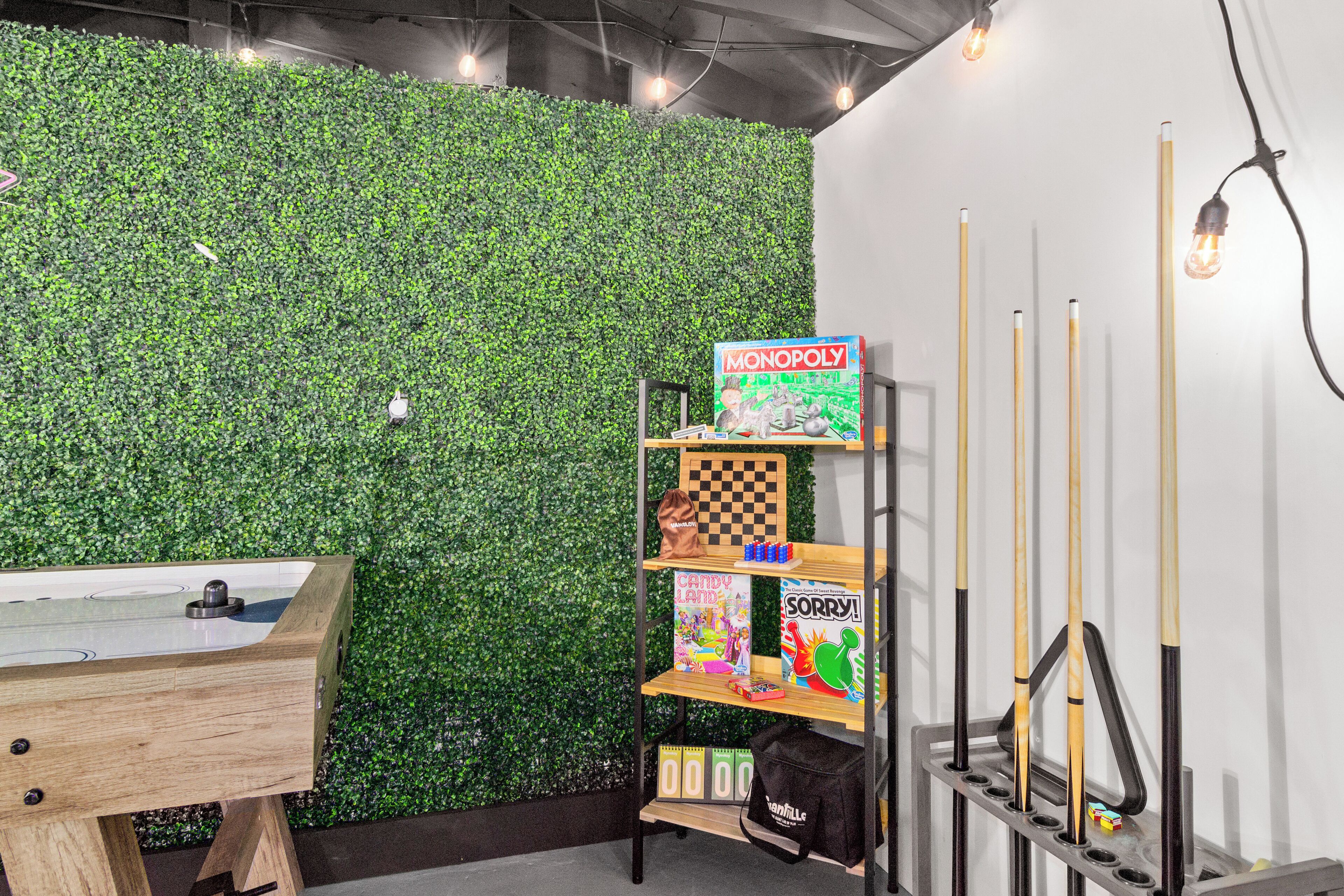 A room with a green wall, a pool table, and shelves with board games and pool cues.