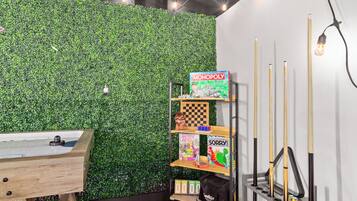 A room with a green wall, a pool table, and shelves with board games and pool cues.