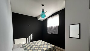 A small, dimly lit room with a single bed, a window with curtains, a wall-mounted mirror, and a ceiling light fixture. - Apartment in front of the sea (Siderno)