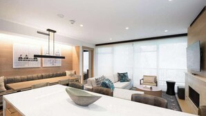 A modern hotel room with a large dining table, a sofa, a TV, and a fireplace.