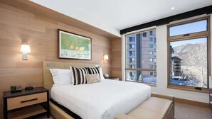 A modern hotel room with a large bed, two bedside tables, a nightstand, and a large window offering a view of a mountain and buildings.