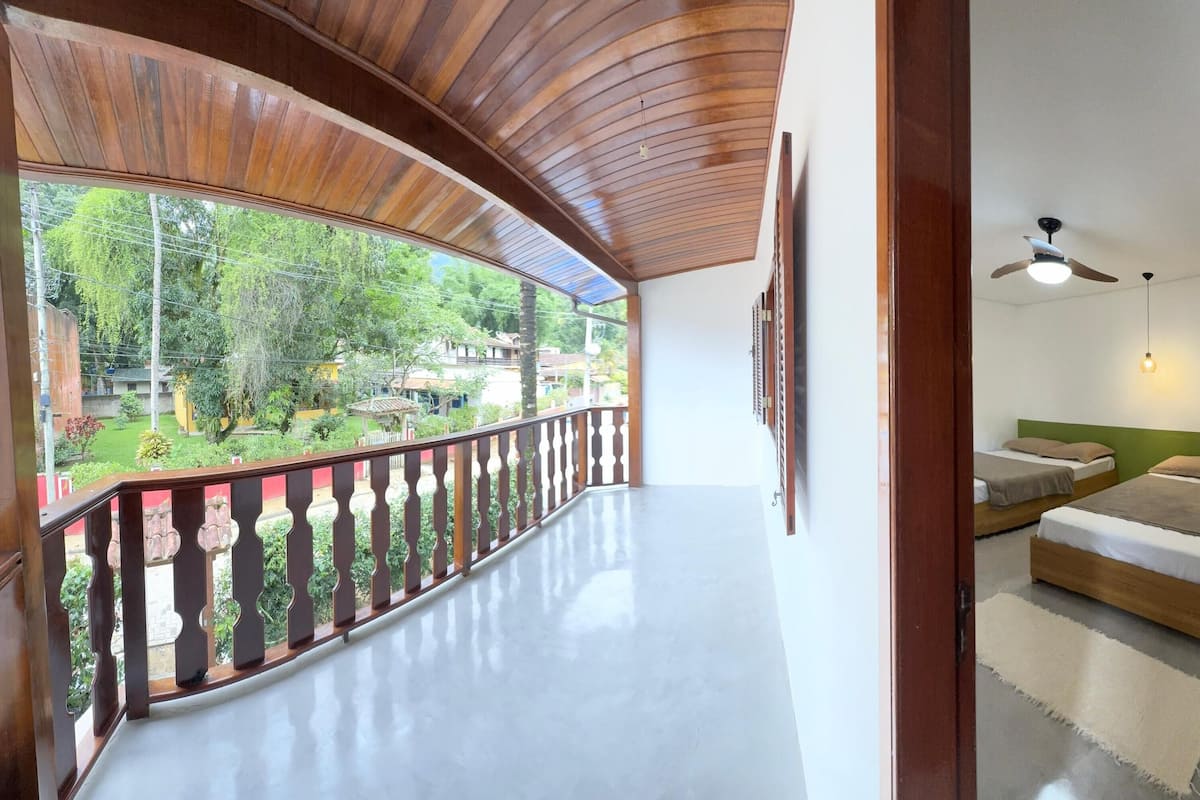 A balcony with a wooden railing overlooking a garden, a bedroom with two beds and a ceiling fan, and a hallway with a door.