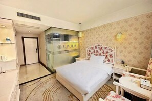 Habitación de hotel con cama, mesita de noche y un pequeño escritorio con silla. Cuenta con un tabique de vidrio y una puerta que da a otra habitación.