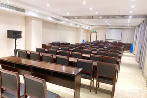 Un salón de conferencias con filas de sillas y mesas, una pantalla de proyector y un orador.