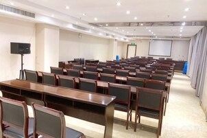 Un salón de conferencias con filas de sillas y mesas, una pantalla de proyector y un orador.