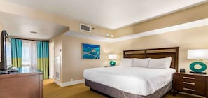 Quarto de hotel com uma cama grande, uma mesa com televisão, uma mesinha de cabeceira com abajur e um quadro na parede.