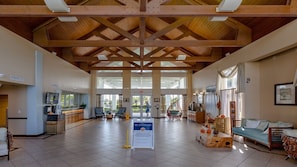 A spacious lobby with a high ceiling, wooden beams, a reception desk, seating area, and a sign indicating no gatherings.