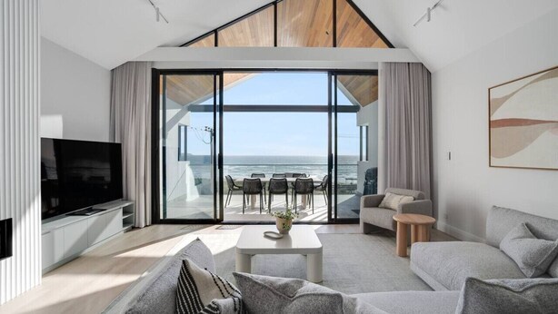 Een moderne woonkamer met een grote televisie, grijze banken, een salontafel en uitzicht op de zee door grote glazen deuren.