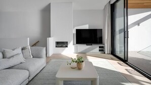 Een moderne woonkamer met een grijze bank, een witte salontafel met een plant, een open haard en een grote televisie.