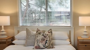 A bedroom with a large bed, two bedside tables with lamps, a window with blinds, and a view of trees outside.