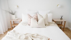 A neatly made bed with white bedding and a few decorative pillows, two bedside tables with lamps, and a small potted plant on the left side. - Casa Billie ~ Family friendly beachside oasis (Aldinga Beach)