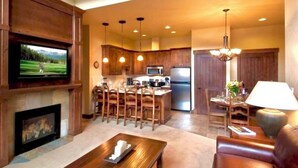 A spacious living room with a fireplace, a television mounted on the wall, a dining area with wooden chairs, and a kitchen with wooden cabinets and a refrigerator. - Grand Lodge Peak 7: True Ski-in Ski-out resort Jan 10-17,2026  Breckenridge, CO  (Breckenridge)