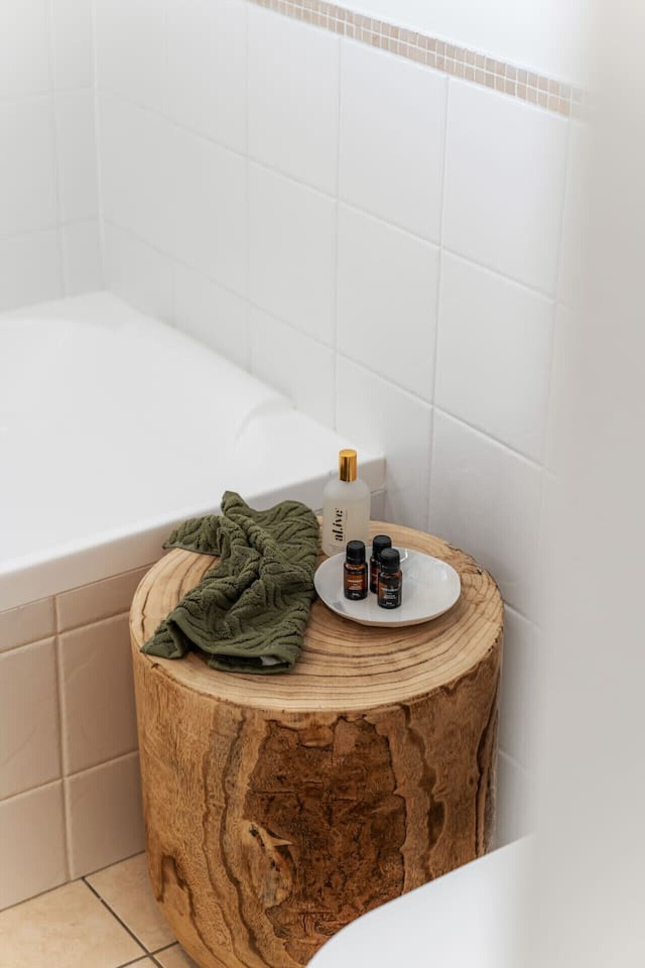 A bathroom with a bathtub, a round wooden stool, a green towel, and three small bottles on a plate.