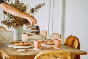 Una mesa de comedor de madera con platos, tazas y una persona sirviendo una bebida.