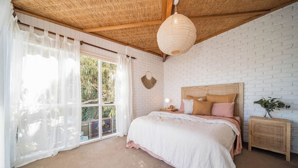 Un dormitorio con cama, mesita de noche, lámpara de pared y ventana con cortinas.