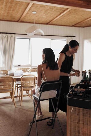 A room with a dining table, chairs, a person sitting, and another person standing by a counter with various items.