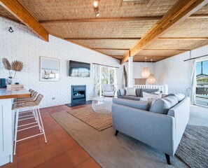 A modern living room with a sofa, a dining area with bar stools, a fireplace, and a large window with a view. - Salt Bush Beach House~The ultimate Fleurieu escape (Aldinga Beach)