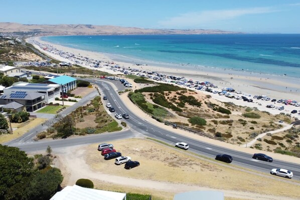 A coastal road with parked cars, a beach with people, and a building with solar panels.