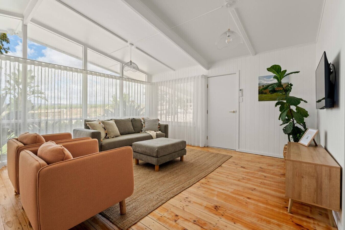 A spacious living room with a large window, a grey sofa set, a brown armchair, and a wooden cabinet.