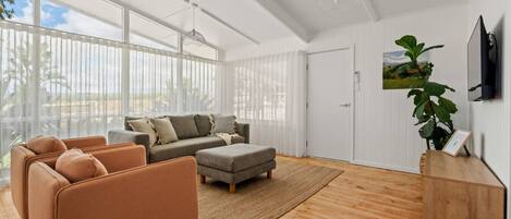 A spacious living room with a large window, a grey sofa set, a brown armchair, and a wooden cabinet.
