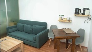 Habitació amb un sofà color turquesa, una taula de fusta i una petita taula de menjador. Hi ha dues cadires, una estantàpia de fusta amb objectes de cuina i una cafetera penjada a la paret.