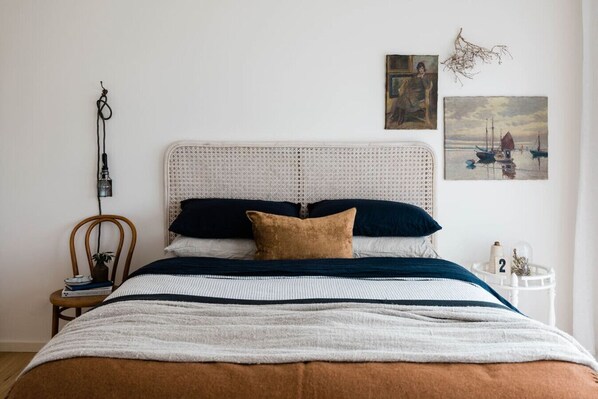 A neatly made bed with a white headboard, a wooden chair, and a small table with books and a lamp. - Seaside surf haven, pup friendly - Seafarers Lodge (Middleton)