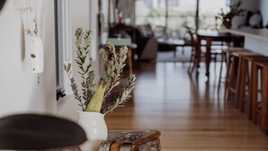 Un couloir avec un coffre en bois rustique, un vase blanc contenant des plantes séchées et un coin repas avec des meubles en bois et des luminaires suspendus.