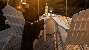 Une terrasse avec deux fauteuils Adirondack, une petite table ronde sur laquelle se trouvent une bouteille et un verre, et une vue sur le coucher de soleil au-dessus d’un paysage vallonné.