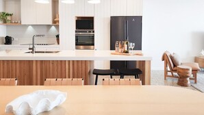 Et moderne køkken med en central ø, indbygget ovn og et sort køleskab. Træstole og et lille træbord med en hvid genstand på.