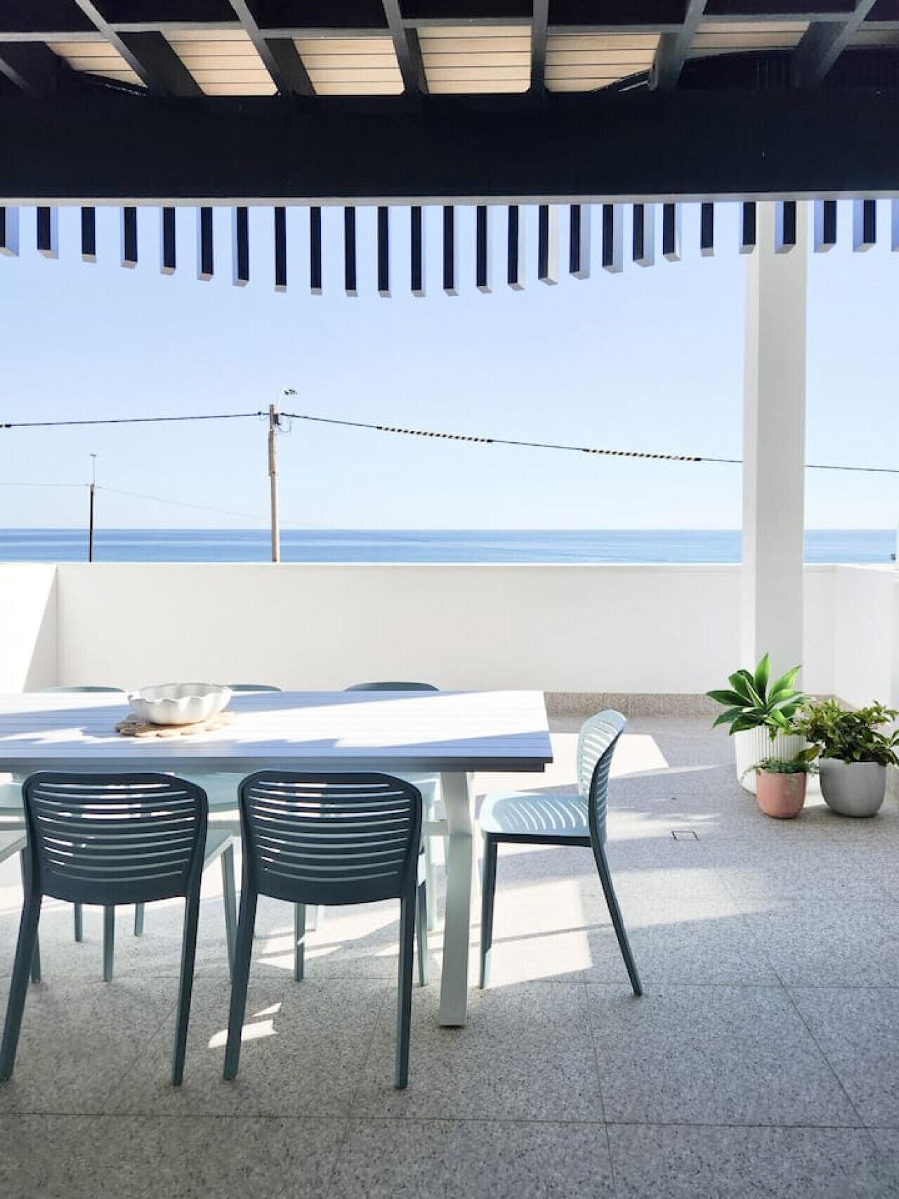 Un patio exterior con mesa y sillas, vista al mar y plantas en macetas.