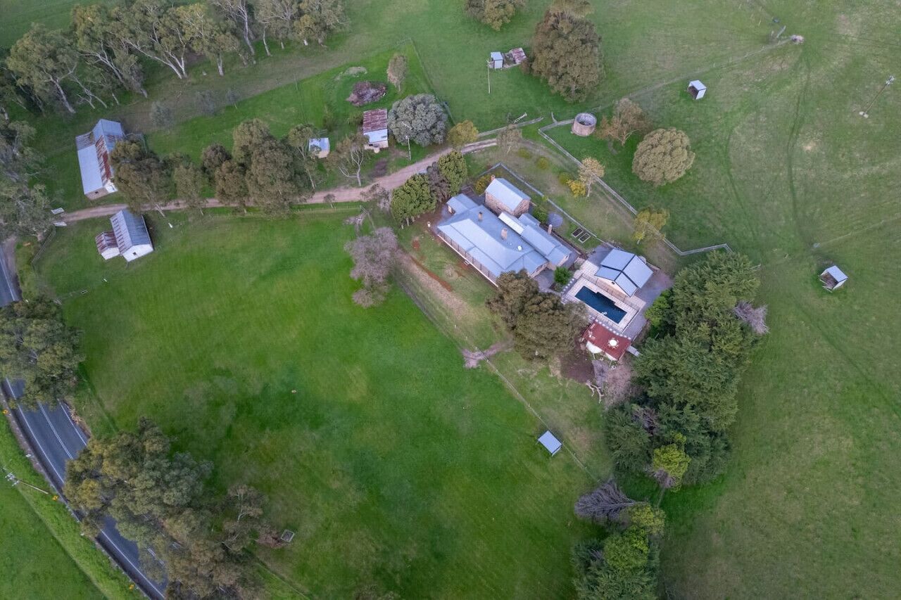 An aerial view of a rural property with multiple buildings, including a main house with a large garage and a swimming pool, surrounded by green fields and scattered trees.