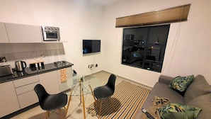 Un espacio habitable compacto con kitchenette, una zona de comedor con dos sillas, un sofá y un ventanal con persianas.