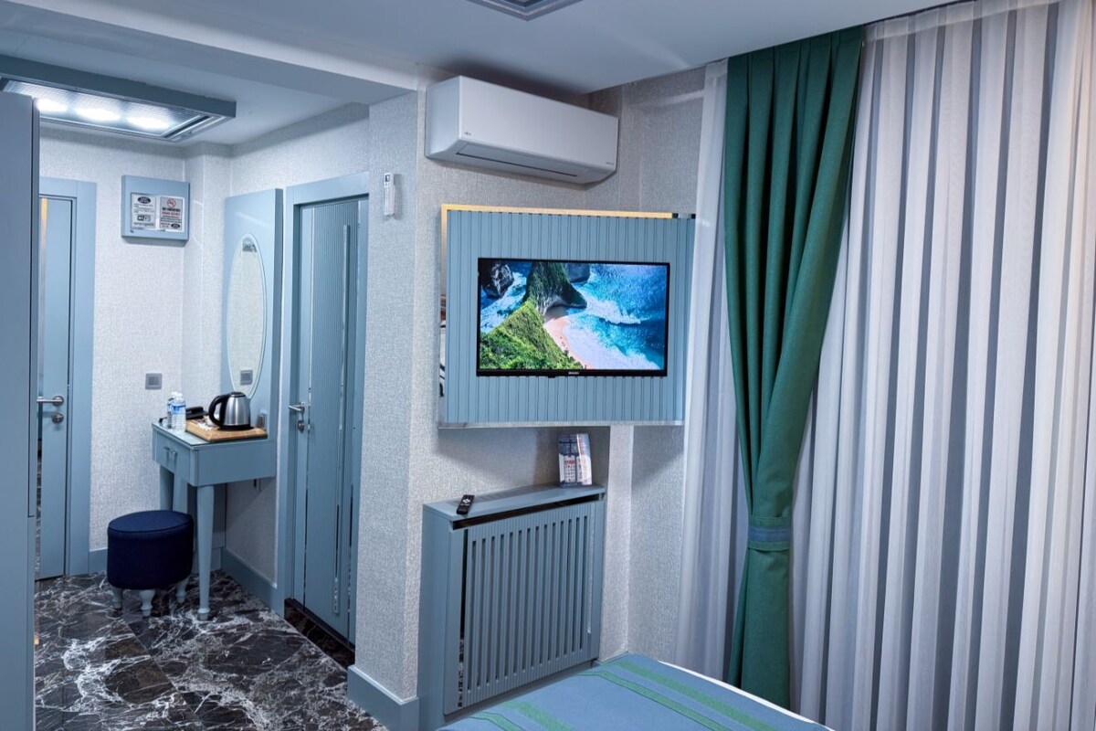 A hotel room with a television mounted on the wall, a small table with a kettle, a mirror, and a radiator.